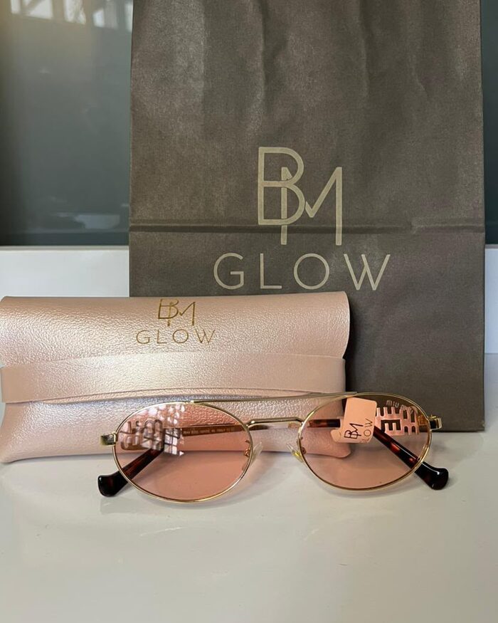 Sacola e estojo rosé da BM GLOW ao lado de um óculos rosé